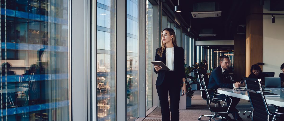 Eine Frau läuft im Büro entlang, während im Hintergrund an einem Verhandlungstisch gearbeitet wird